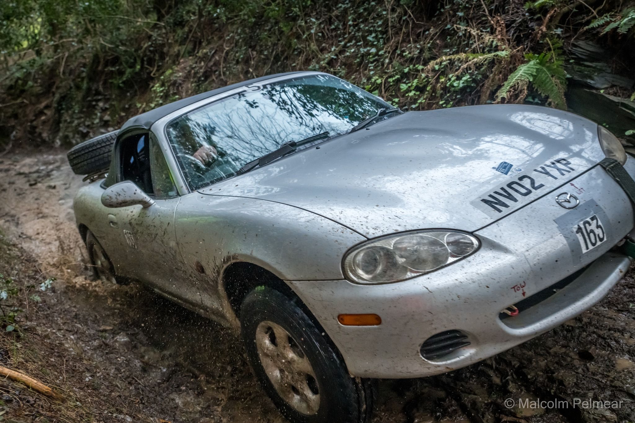 Mud-splattered silver Mazda MX-5 rally car traversing a challenging off-road trial track with a spare tire visible.