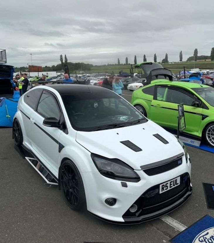 White modified Ford Focus RS Mk2, owned by Mark George, with a black roof and black wheels, on display next to a bright green Focus at an outdoor car show.