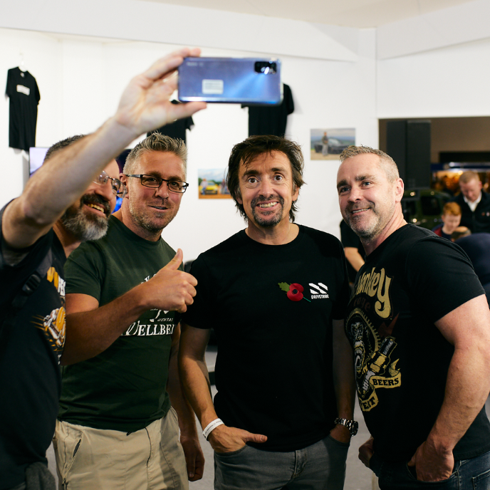 Richard Hammond posing for a fan selfie with three male fans at an event. Hammond is wearing a black t-shirt with a poppy and "DRIVETRIBE" logo.