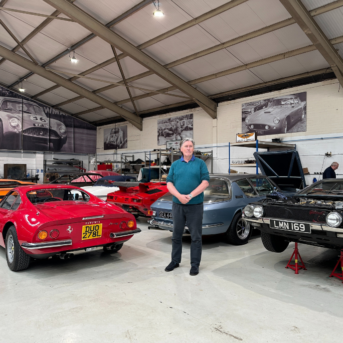 The owner of Tyrrell's Classic Workshop standing in the middle of a garage surrounded by a collection of classic cars, including a red Ferrari Dino and a black Jensen Interceptor.