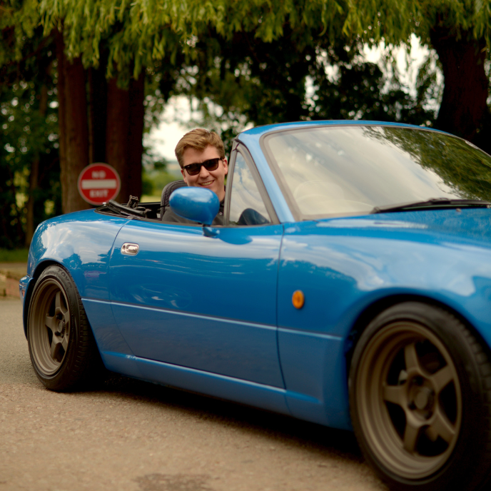 Luke from Garage of Luke driving and smiling in a bright blue convertible Mazda MX-5 Miata with aftermarket wheels, parked on a road lined with large trees.