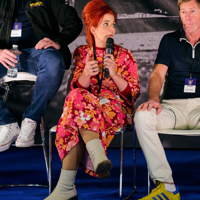 idriveaclassic, a woman with vibrant red hair, speaking into a microphone while sitting on a panel, wearing a striking pink and orange floral print dress and white boots.