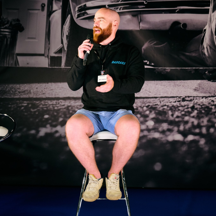 	DadCars founder, a man with a red beard, speaking into a microphone while sitting on a stool at a car event. He wears a black "DADCARS" hoodie and blue shorts.