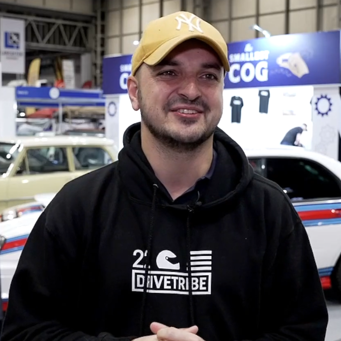 Mike Fernie from DRIVETRIBE wearing a black hoodie with the DRIVETRIBE logo and a yellow New York Yankees baseball cap, smiling in front of an event booth.