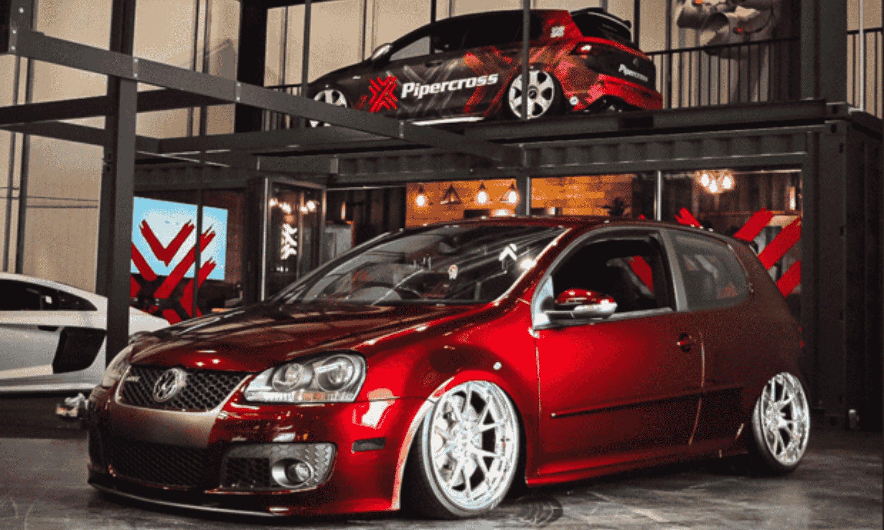 Low-rider red Volkswagen Golf GTI Mk5 with deep dish wheels on display beneath a black and red Pipercross branded performance hatchback raised on a metal platform at an indoor car show.