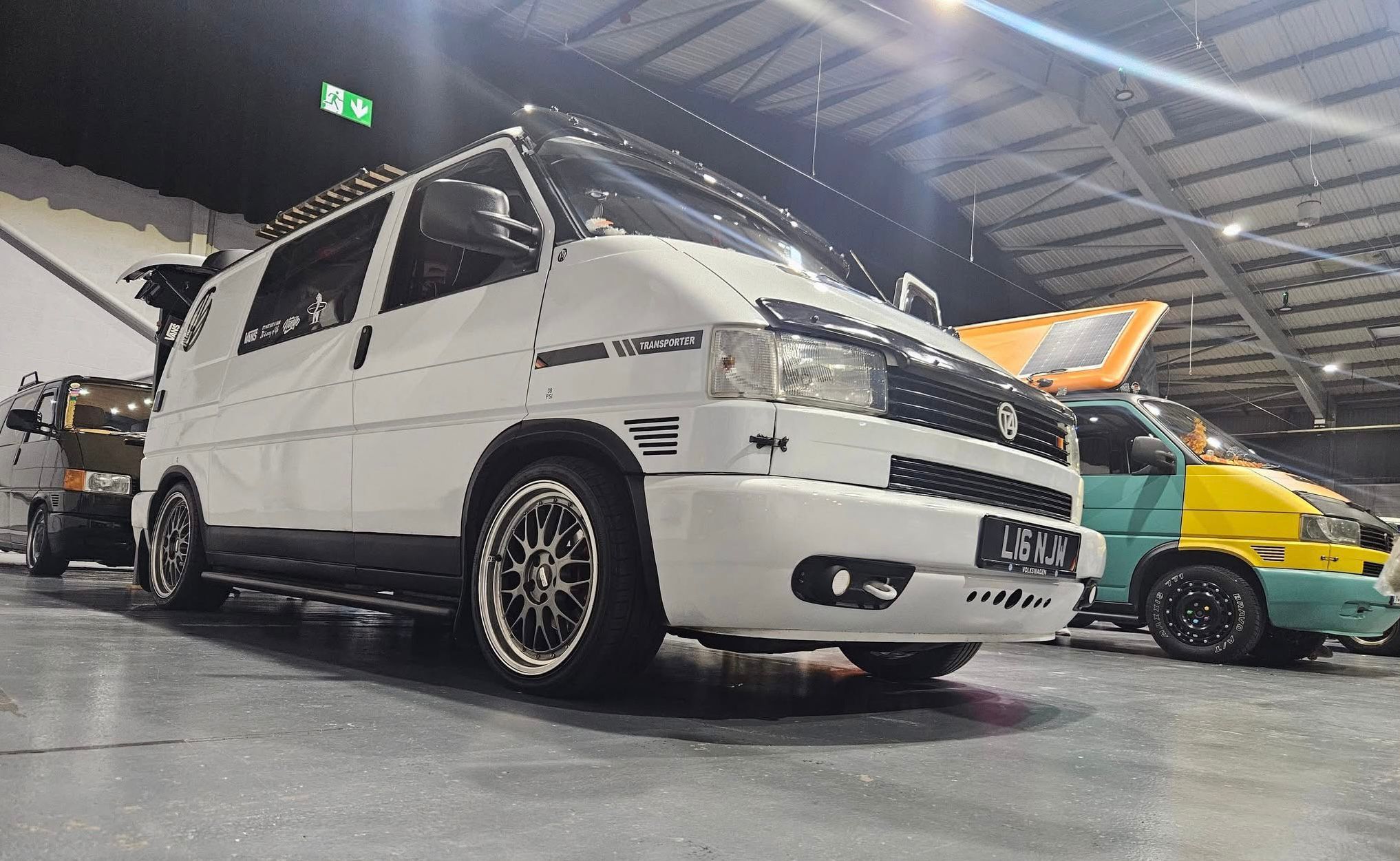 A white modified, lowered Volkswagen T4 Transporter van, owned by Nigel Walford, with aftermarket wheels and a roof rack, parked indoors at an auto show.