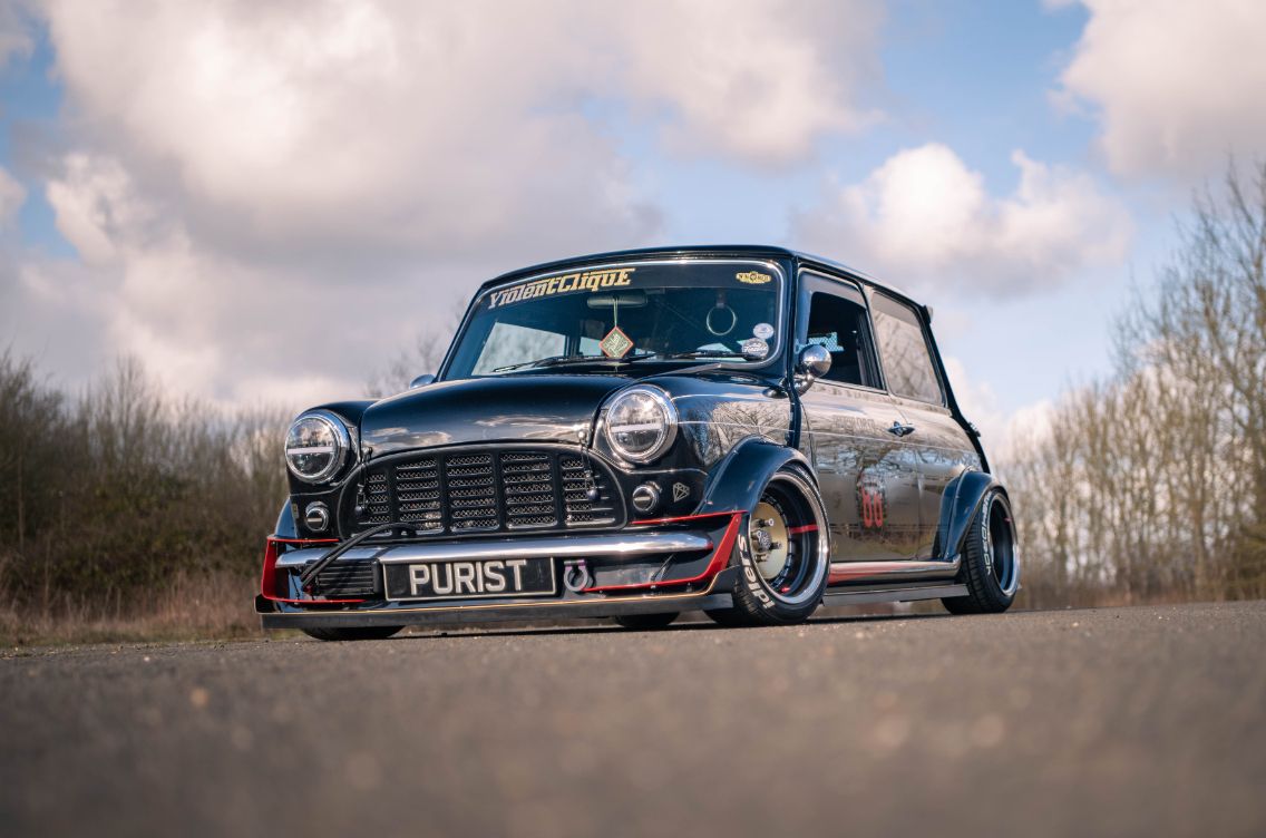 Stanced black classic Classic Mini with front oil cooler, custom body kit, and PURIST license plate parked outdoors, shot by Well Chuffed Media.