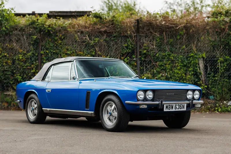 Vintage blue Jensen Interceptor convertible parked on pavement with a brick wall behind it.