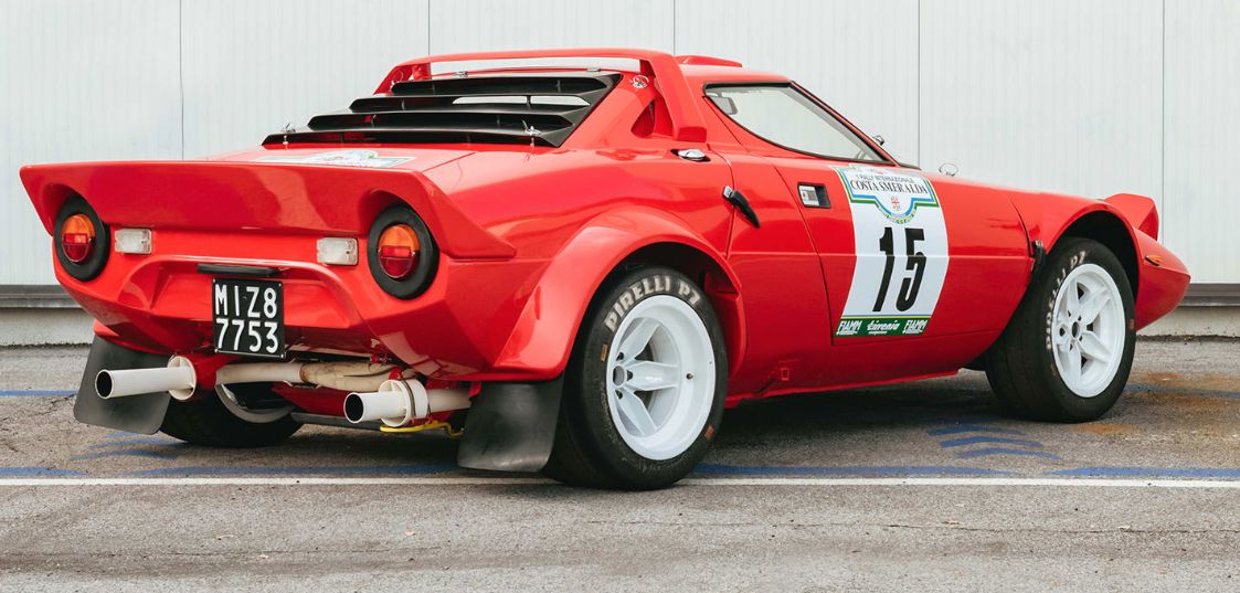 Red Lancia Stratos rally car with white wheels and racing number 15 parked against a white wall.