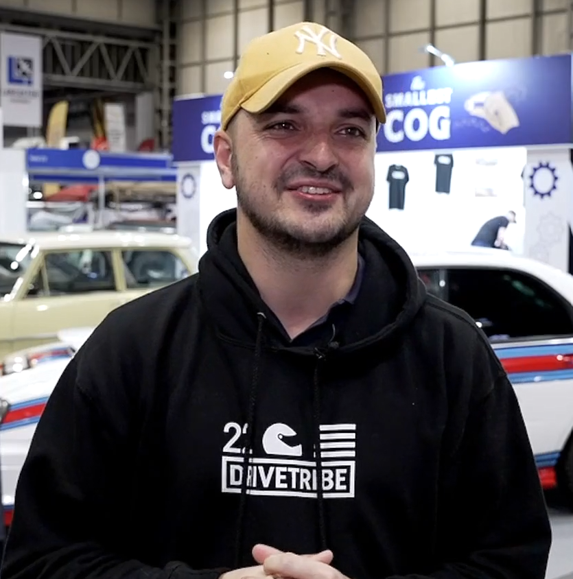  Mike Fernie, a presenter for DRIVETRIBE, is smiling at an automotive event while wearing a branded hoodie and a baseball cap.