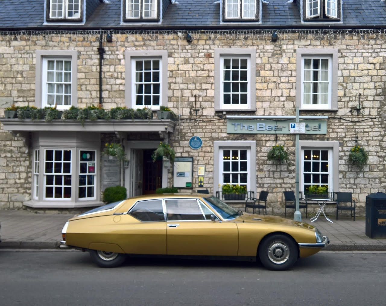 A side profile of a gold Citroën SM grand tourer, owned by Steven Prevett, parked on a street in front of a stone building with the sign 'The Bear Hotel'.