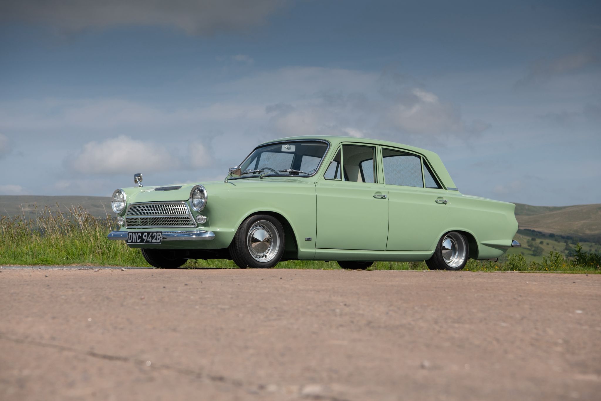 A pristine light green classic Ford Cortina Mark I sedan, owned by Will Wilson, with chrome trim, parked on a road against a backdrop of rolling green hills.