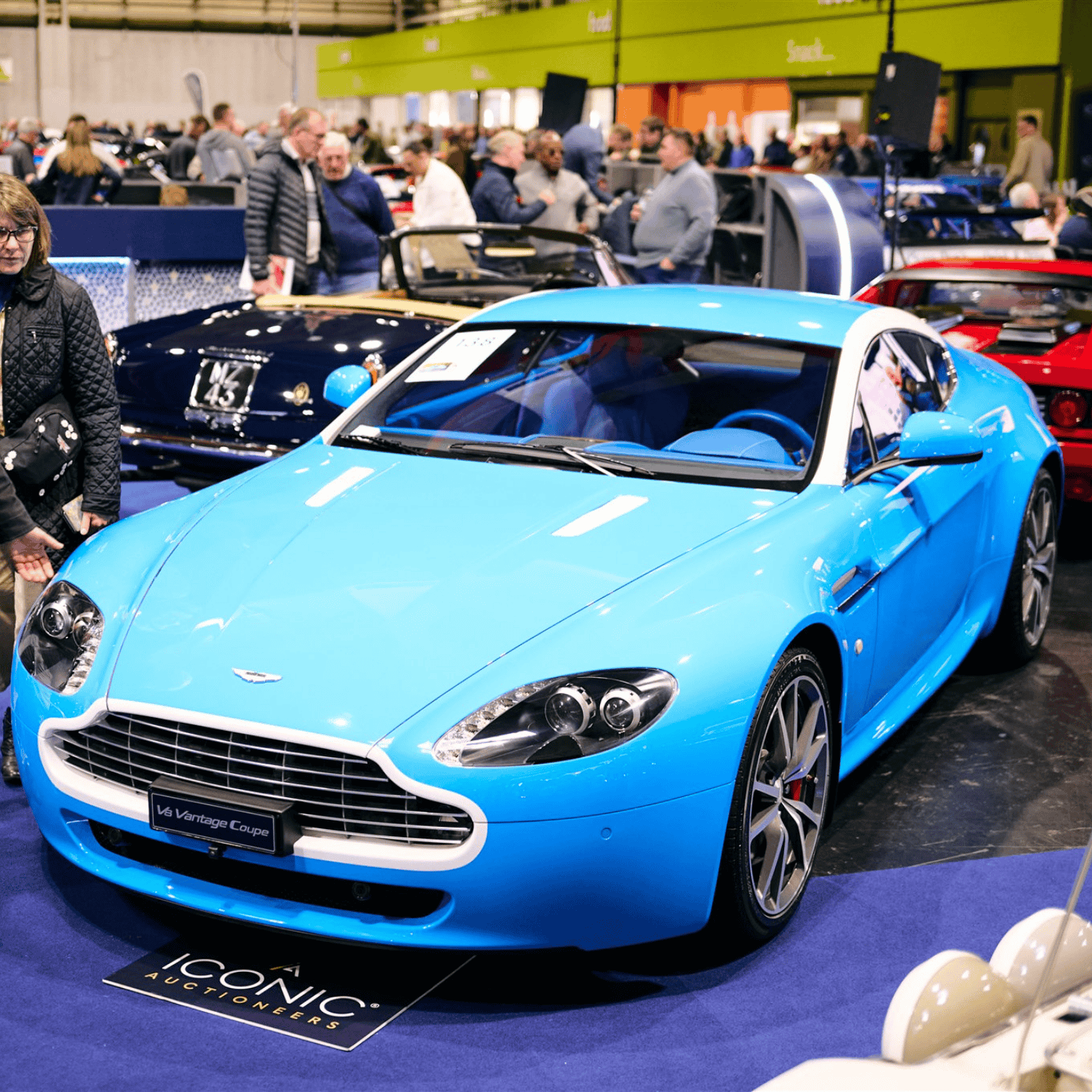 Bright blue Aston Martin V8 Vantage Coupe on display at a classic car show. The sleek sports car is in the foreground, with other luxury vehicles and a crowd of car enthusiasts visible in the background.