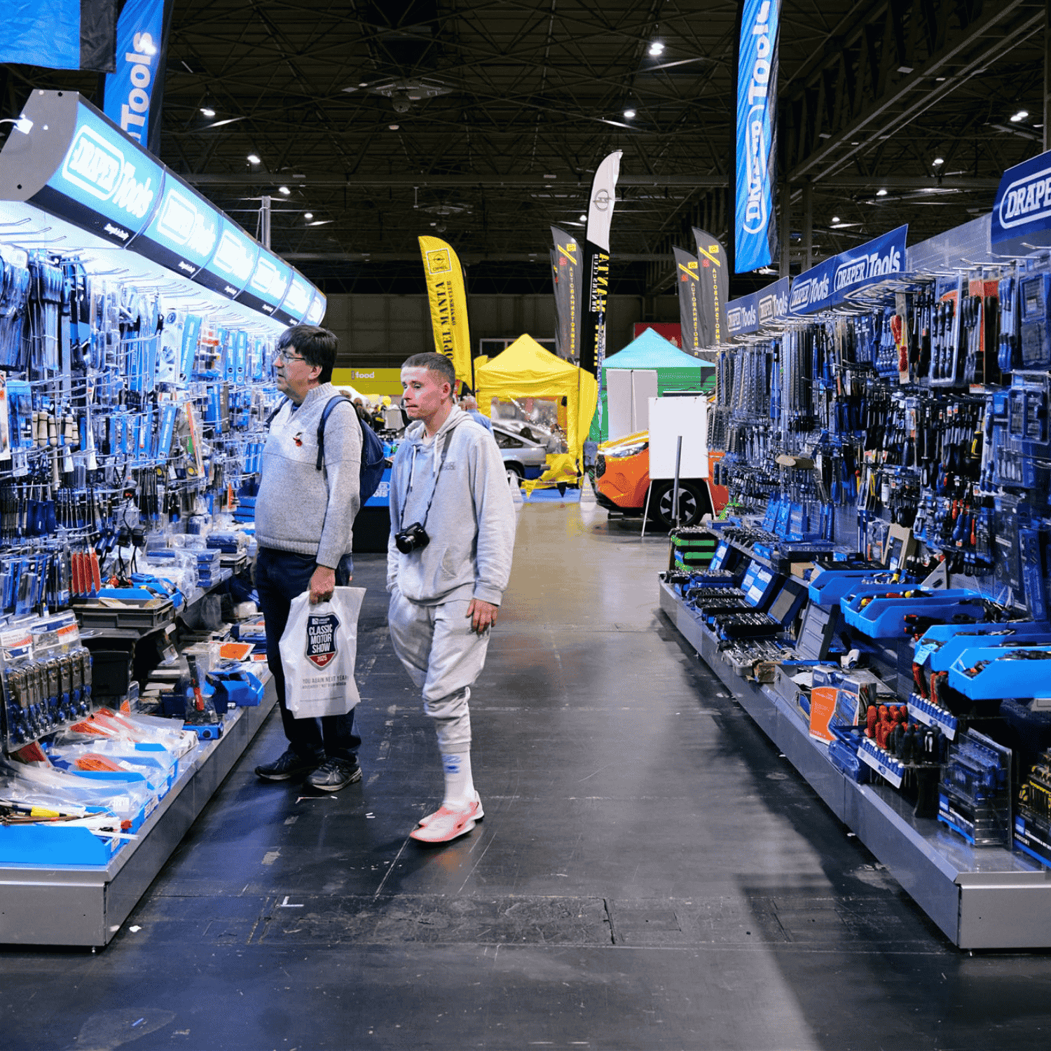 Two people browsing an aisle of tools and automotive parts at a large trade show. The men are looking at the shelves filled with various items, with banners for brands like "Draper Tools" overhead.