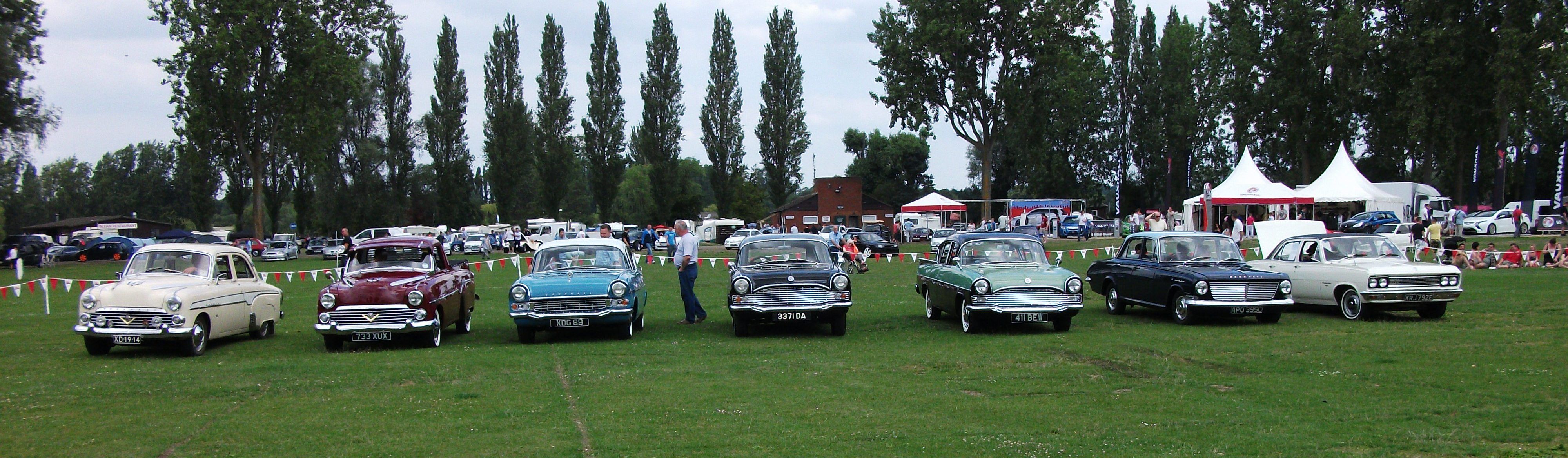 A wide shot of a grassy field with a line of classic cars. The cars are arranged from left to right, with a light-coloured Austin Cambridge at the far left and five different generations of Vauxhall Victor sedans in various colours, including red, light blue, dark blue, and white. In the background, there are trees, a brick building, and a series of white tents, suggesting a car show or outdoor event.