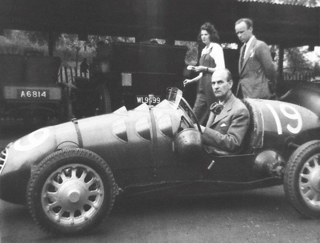 A man in a race car with a woman and another man standing behind him, vintage photo.