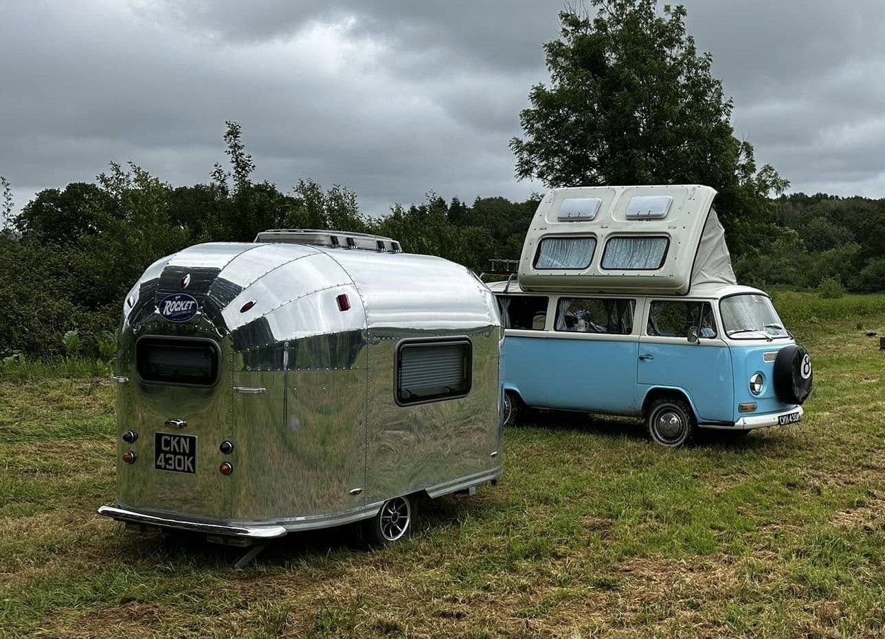 Vintage blue VW camper van and a shiny silver Airstream-style trailer parked in a field.