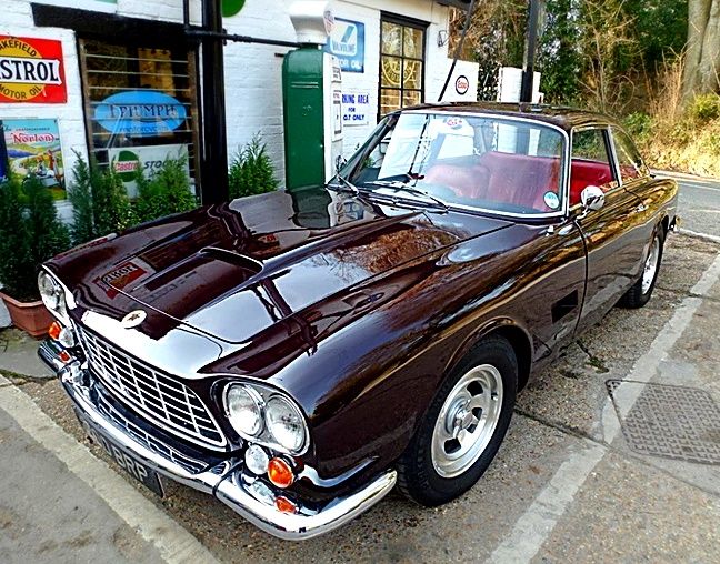A classic dark maroon Gordon-Keeble GK1 coupe with a red leather interior, parked in front of a vintage-style petrol station with retro Wakefield and Castrol motor oil signs.