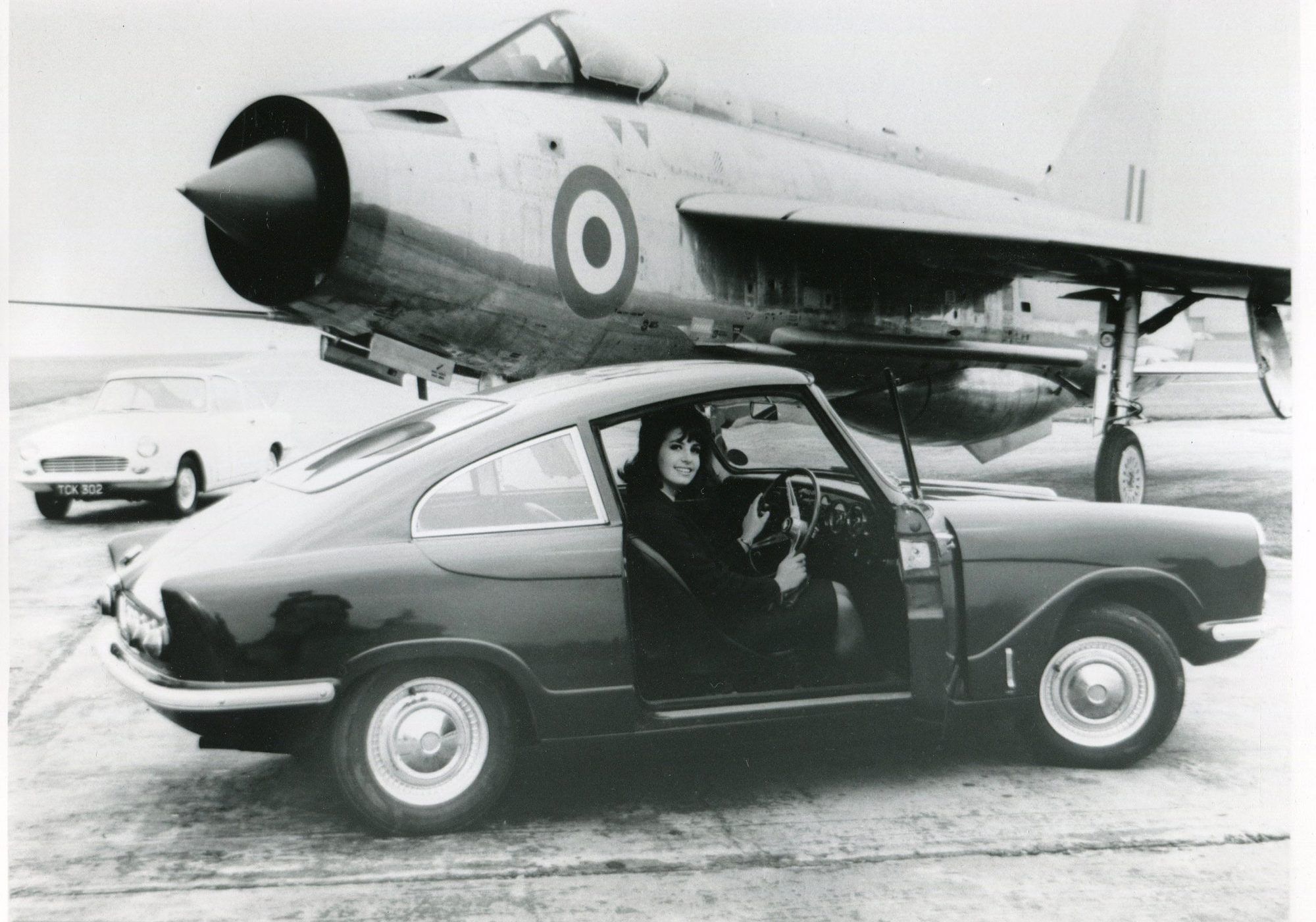 Black and white photo of a woman in a Bond Triumph Spitfire Mk3 convertible parked in front of an English Electric Lightning jet fighter.