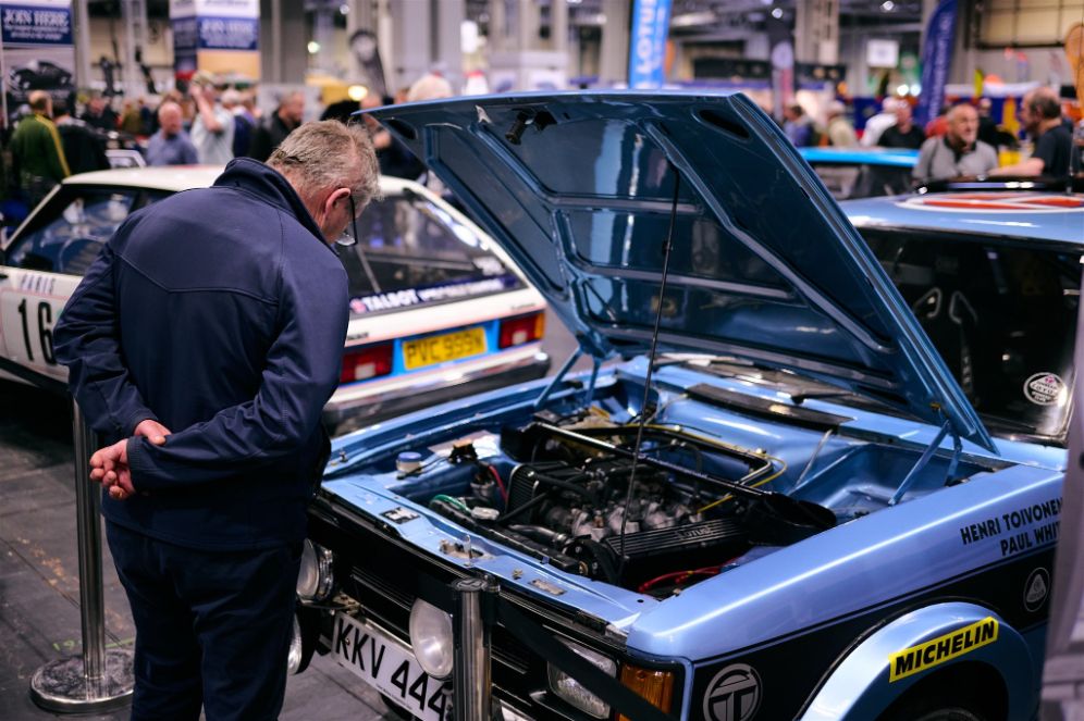 A visitor examining the engine of a blue classic rally car, possibly a Talbot Sunbeam Lotus tribute to Henri Toivonen, at the Classic Motor Show.