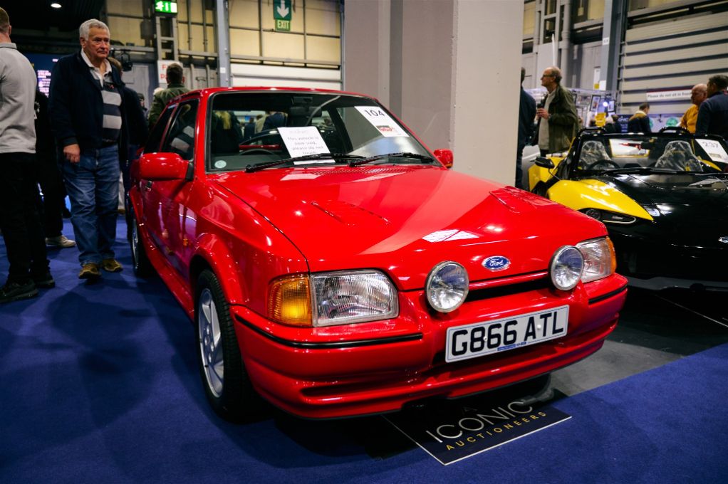 A bright red Ford Escort RS Cosworth on display at the Classic Motor Show, with an Iconic Auctioneers sign nearby.