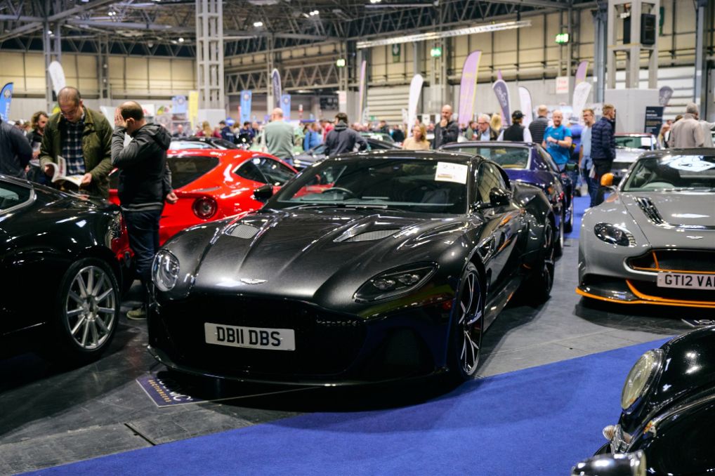 Crowded classic car show floor showcasing modern Aston Martin sports cars including a black DBS and a grey GT12, with other classic cars and attendees.