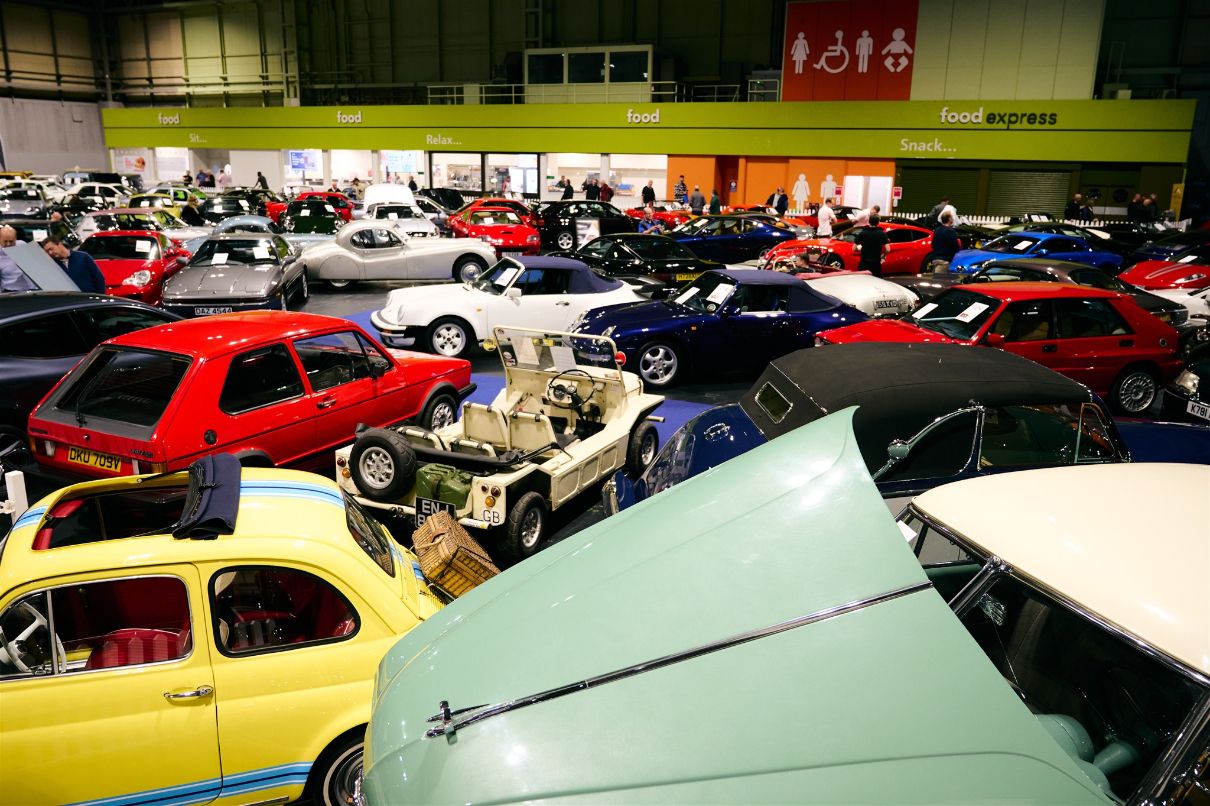 Crowded indoor classic car show display featuring a wide variety of vintage cars including sportscars, saloons, and a cut-away chassis, with food vendor signs overhead.