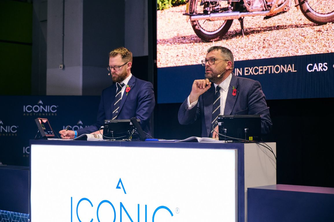 Auctioneers from Iconic Auctioneers at a classic car show, conducting a live auction event with a large screen displaying cars.
