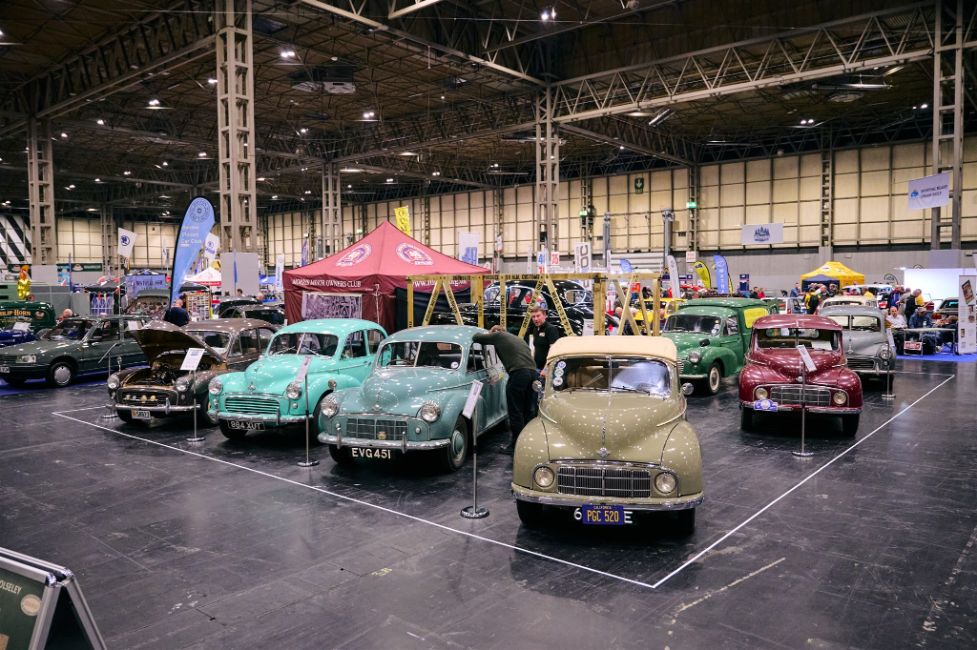 A line of diverse classic cars, including various saloon models in shades of green, blue, and beige, displayed indoors at a spacious motor show.