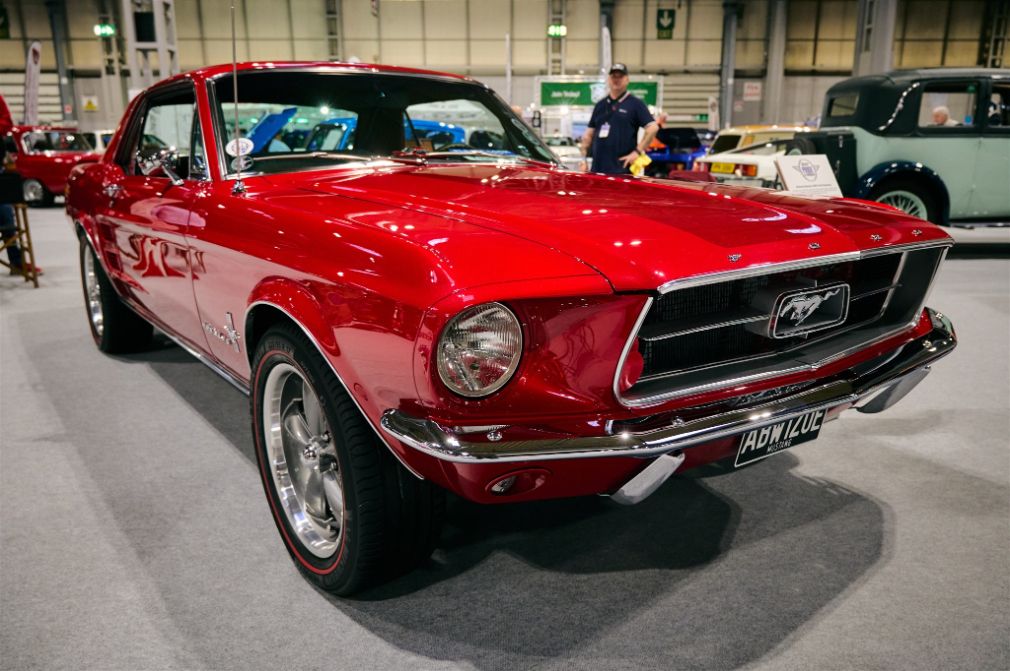 A striking red Ford Mustang is the main subject of this image, captured at an indoor motor show. The vintage American muscle car is presented from a low-angle perspective, highlighting its shiny paint, chrome bumpers, and classic styling. The license plate on the front of the car reads 