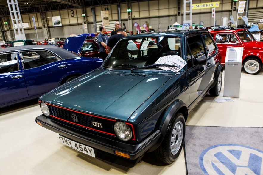 A classic, dark green Volkswagen Golf GTI from the 1980s is on display at a motor show. The car, which is in excellent condition, is the main focus of the image and is positioned at a slight angle. The iconic red stripe on the grille and the GTI badge are clearly visible. In the background, other classic cars, including a red Fiat 500 and a blue coupe, are partially visible. People are also present in the background, walking around and observing the vehicles.
