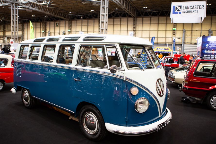 A meticulously restored, two-tone blue and white Volkswagen Type 2 Deluxe from 1964 is on display at an indoor car show. The vintage VW bus, also known as a campervan or microbus, is shown from a side-angle view, highlighting its chrome accents, including the iconic front VW badge and hubcaps. The vehicle has its license plate visible, reading 