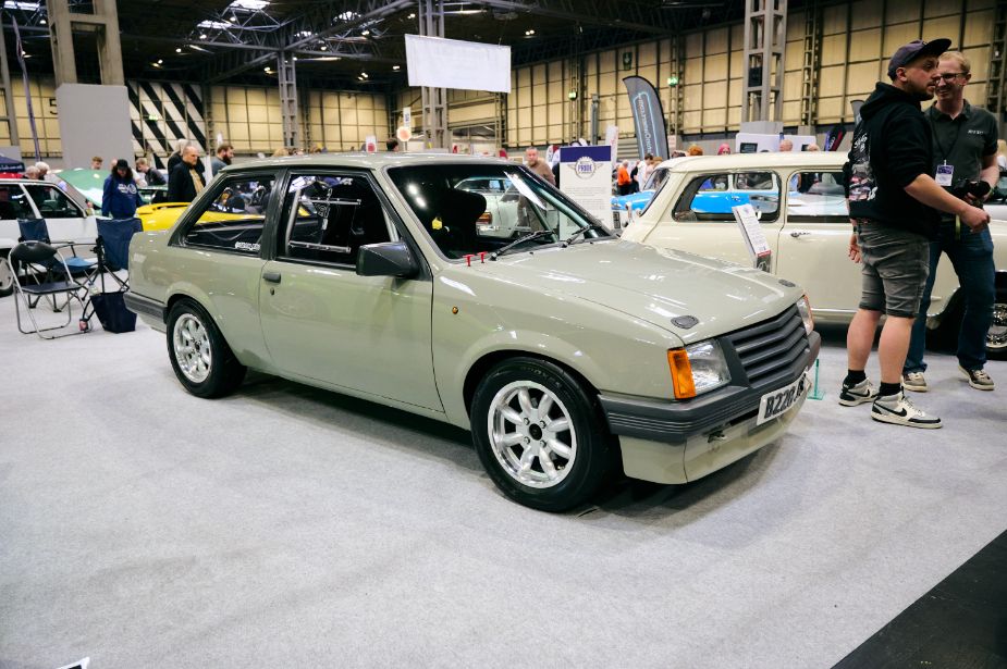 A classic, light gray Vauxhall Nova SR is the main subject of this image, showcased at an indoor car show. The vintage hot hatch is in excellent condition and is viewed from a side angle, highlighting its clean lines, sporty stance, and multi-spoke alloy wheels. A license plate is visible on the front, reading 