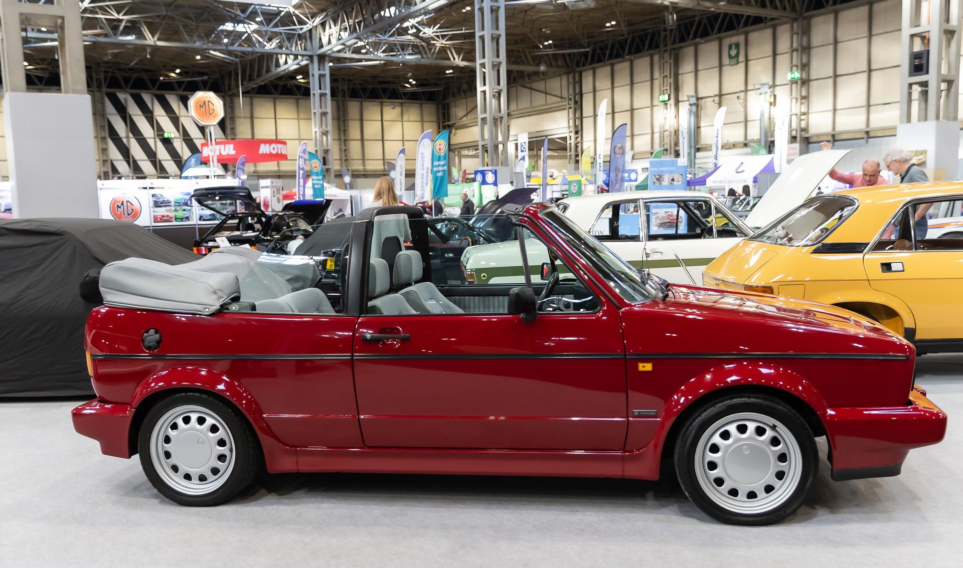 A vibrant red Volkswagen Golf GTI Cabriolet is shown from the side, parked at an indoor classic car show. The soft top is down, revealing the light gray interior. The vintage car is in pristine condition, highlighting its iconic sporty design and clean lines. In the background, other classic vehicles, including a yellow car and a white car with a green stripe, along with event banners and attendees, create a busy show atmosphere.