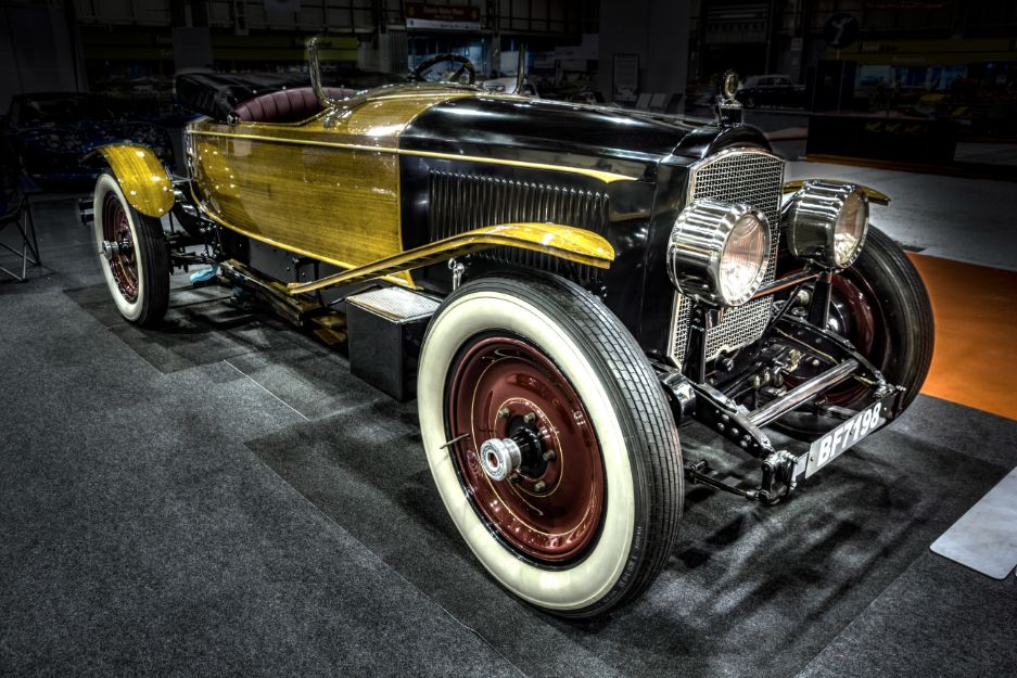  A vintage black and gold Packard car on display at the NEC Classic Motor Show. The car features polished wooden side panels, large headlights, and classic whitewall tires.