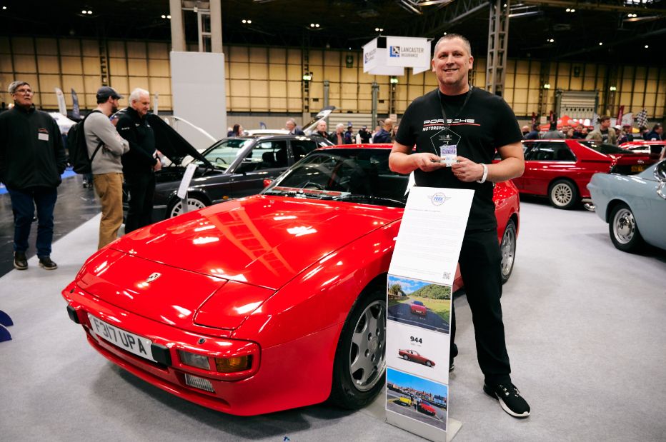 A proud man stands beside a striking red Porsche 944 at an indoor car show. He is holding a trophy and is wearing a black T-shirt with the Porsche Motorsport logo. The classic sports car is in excellent condition, with its front license plate 