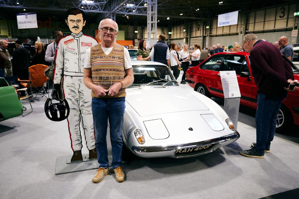 A man is standing next to a vintage, silver-white Lotus Europa sports car at an indoor car show. He is holding his hands clasped in front of him and is positioned directly behind a cardboard cutout of a race car driver. The classic British car is the main subject, with its sleek design and the license plate 