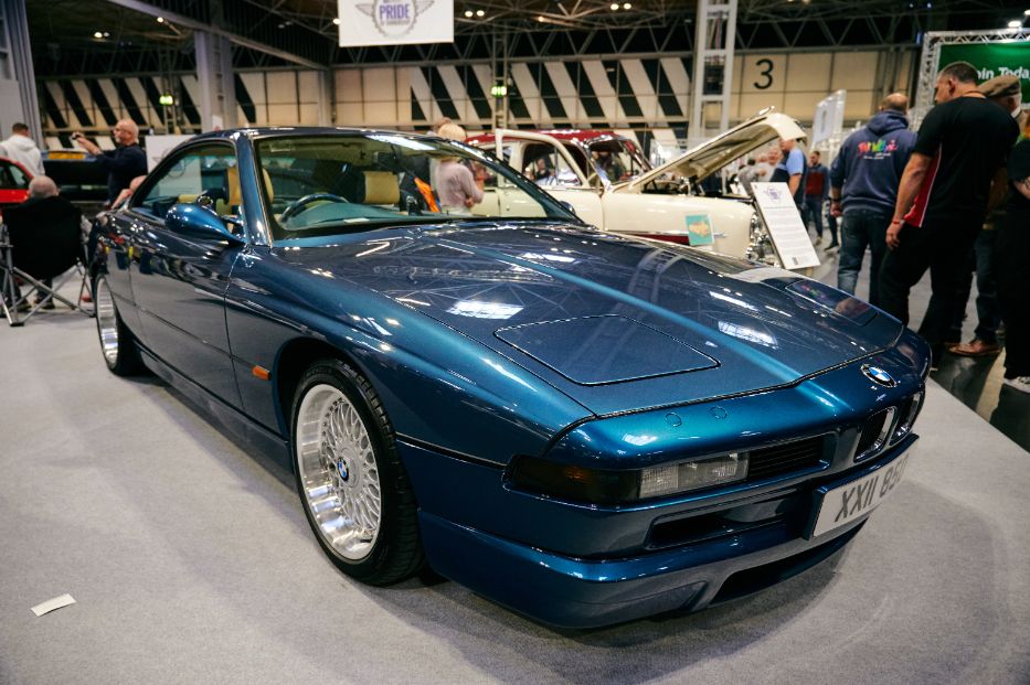 A stunning blue BMW E31 8 Series coupe is the main subject of this image, showcased at an indoor car show. The vintage German grand tourer is in immaculate condition, highlighting its sleek, low-slung design and pop-up headlights. The car features multi-spoke alloy wheels, and its front license plate reads 