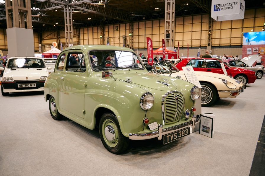A pale green Austin A35 sedan is the main subject of this image, showcased at an indoor classic car show. The vintage car is presented from a head-on angle, highlighting its rounded body, chrome grille, and classic headlights. Its license plate reads 