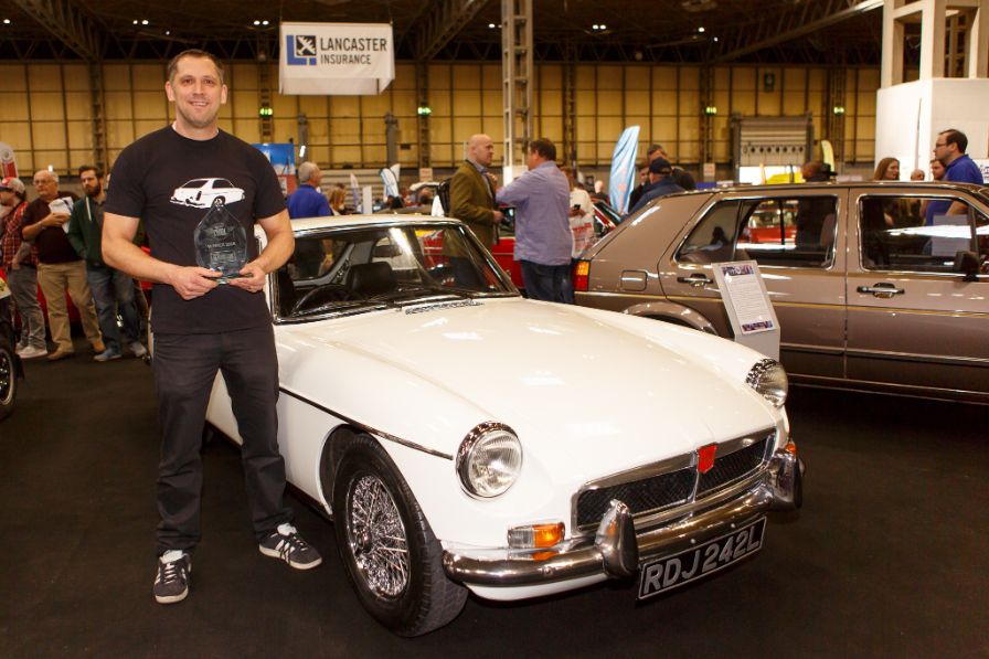 A smiling man in a black T-shirt and dark pants stands next to a pristine white MGB GT convertible at an indoor classic car show. He is holding a clear, glass trophy. The vintage British roadster is the focal point of the image, showcasing its chrome bumper and wire wheels. The license plate on the car reads 