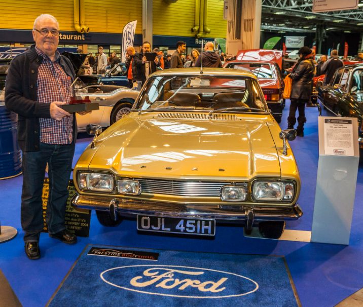 A man, Stuart Turp, stands proudly next to a gold classic Ford Capri at the NEC Classic Motor Show. A Ford logo is on the carpet in front of the car, which appears to be part of a display.