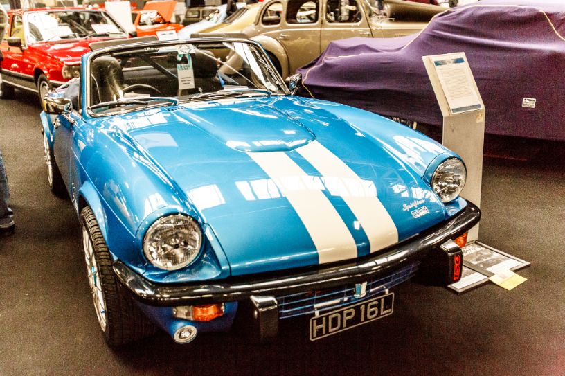 A vintage blue Triumph Spitfire with bold white racing stripes is the main focus of this image, captured indoors at a classic car show. The convertible is meticulously detailed and presented from a head-on, low-angle perspective. The car's chrome bumper and headlights are visible, and its front license plate reads 