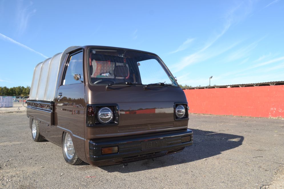 A custom-modified, low-riding brown Kei truck with a canvas cover over its bed is parked on an asphalt lot. The truck has been customized with a dropped suspension, aftermarket chrome wheels, and modern halo headlights. In the background, there is a vibrant red wall and a blue sky with thin clouds. The image is taken at a low angle, highlighting the truck's unique stance and modifications.