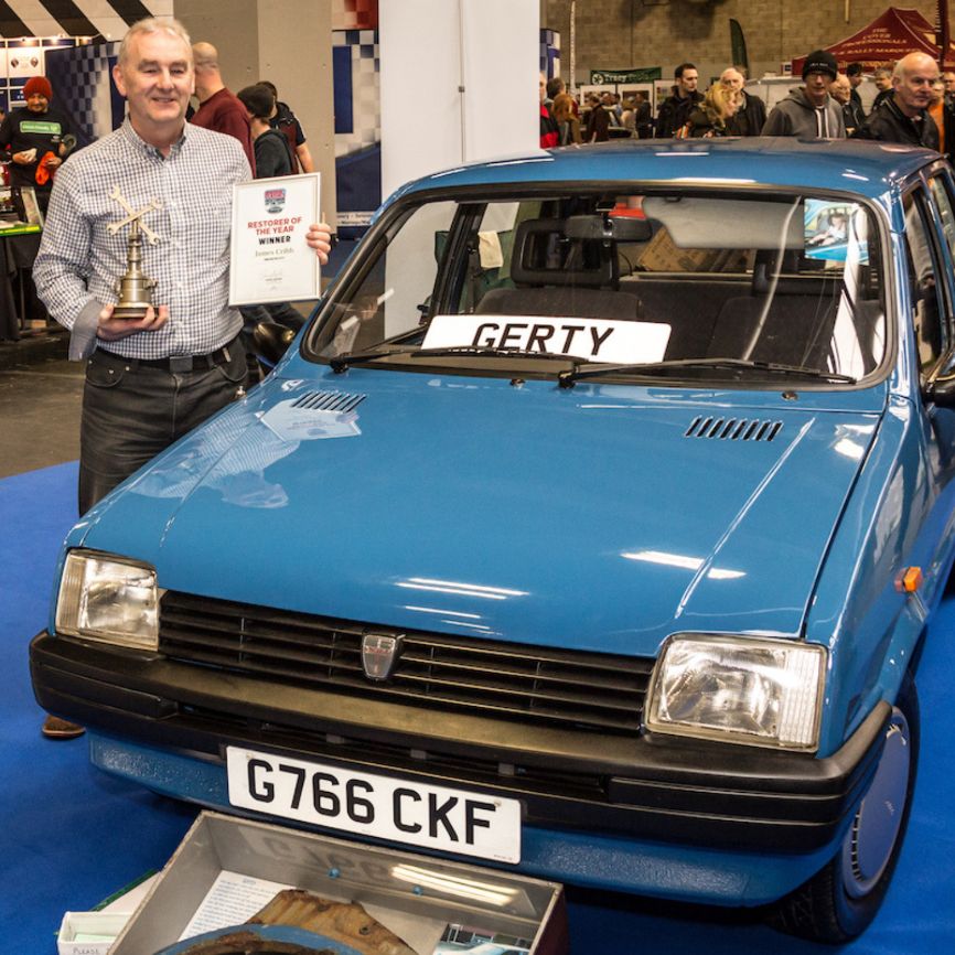 A proud man holds a trophy and a framed certificate, standing next to a classic blue Austin Metro City at an indoor motor show. The man is smiling at the camera, and the car's license plate 
