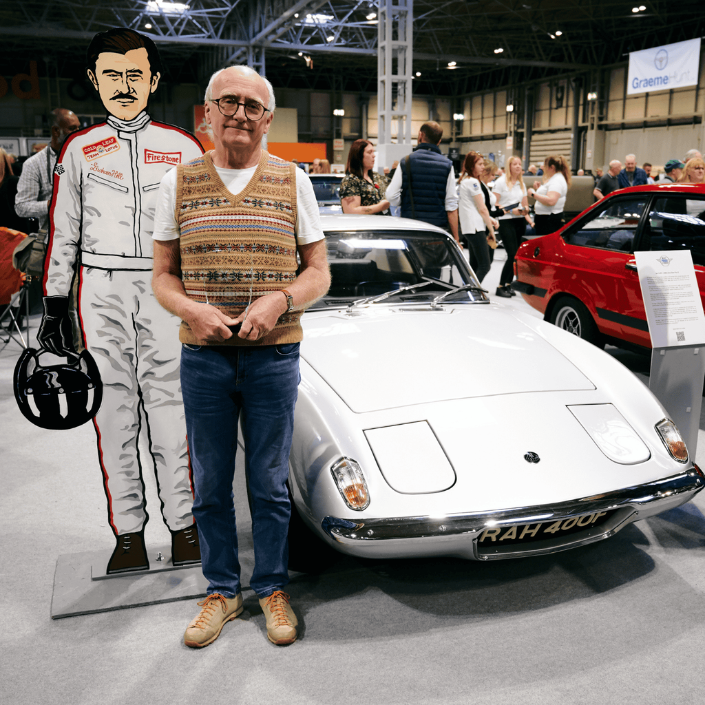 A man is standing next to a vintage, silver-white Lotus Europa sports car at an indoor car show. He is holding his hands clasped in front of him and is positioned directly behind a cardboard cutout of a race car driver. The classic British car is the main subject, with its sleek design and the license plate 