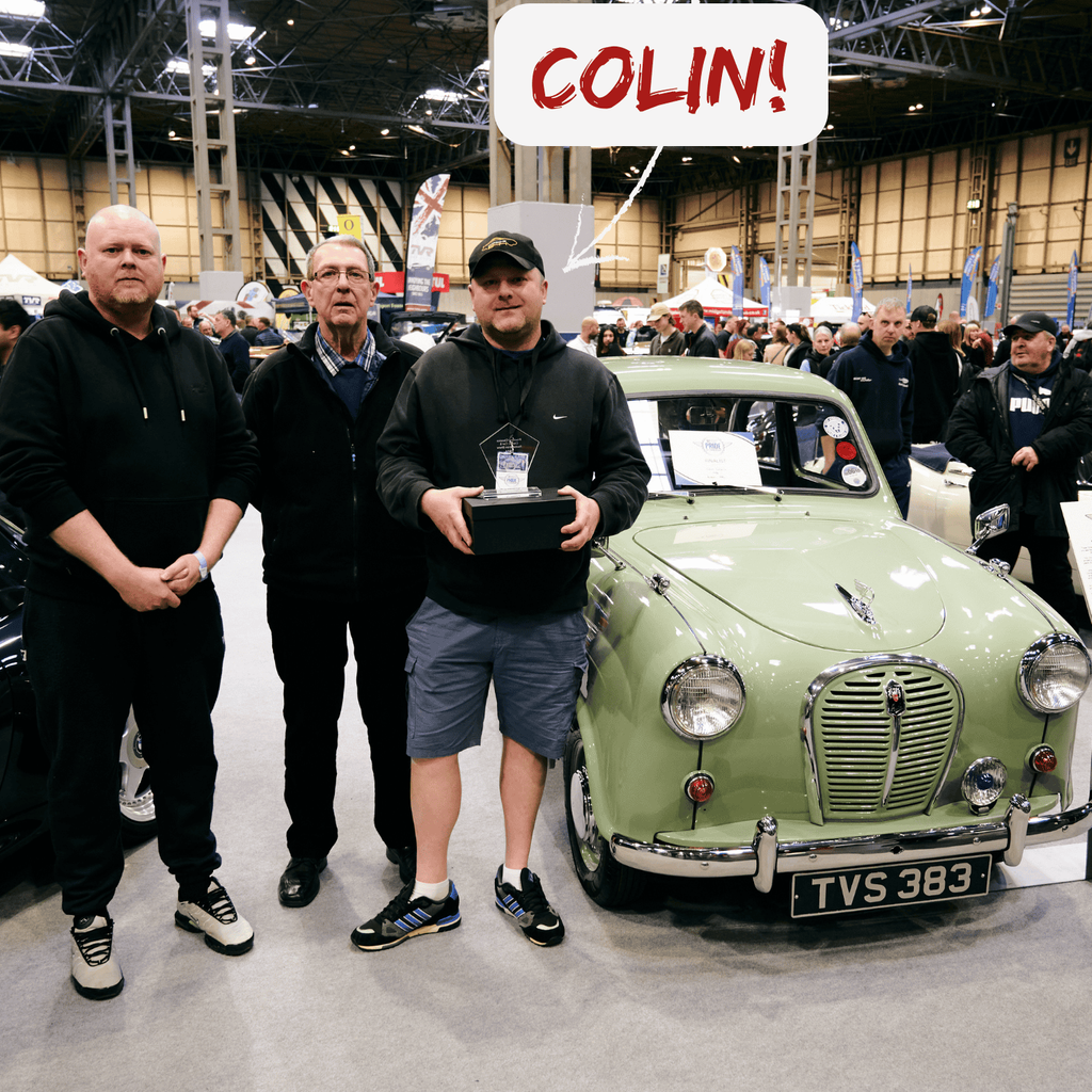 A pale green 1956 Austin A35 sedan is the main subject of this image, showcased at an indoor classic car show. The vintage car is presented from a head-on angle, highlighting its rounded body, chrome grille, and classic headlights. Its license plate reads 