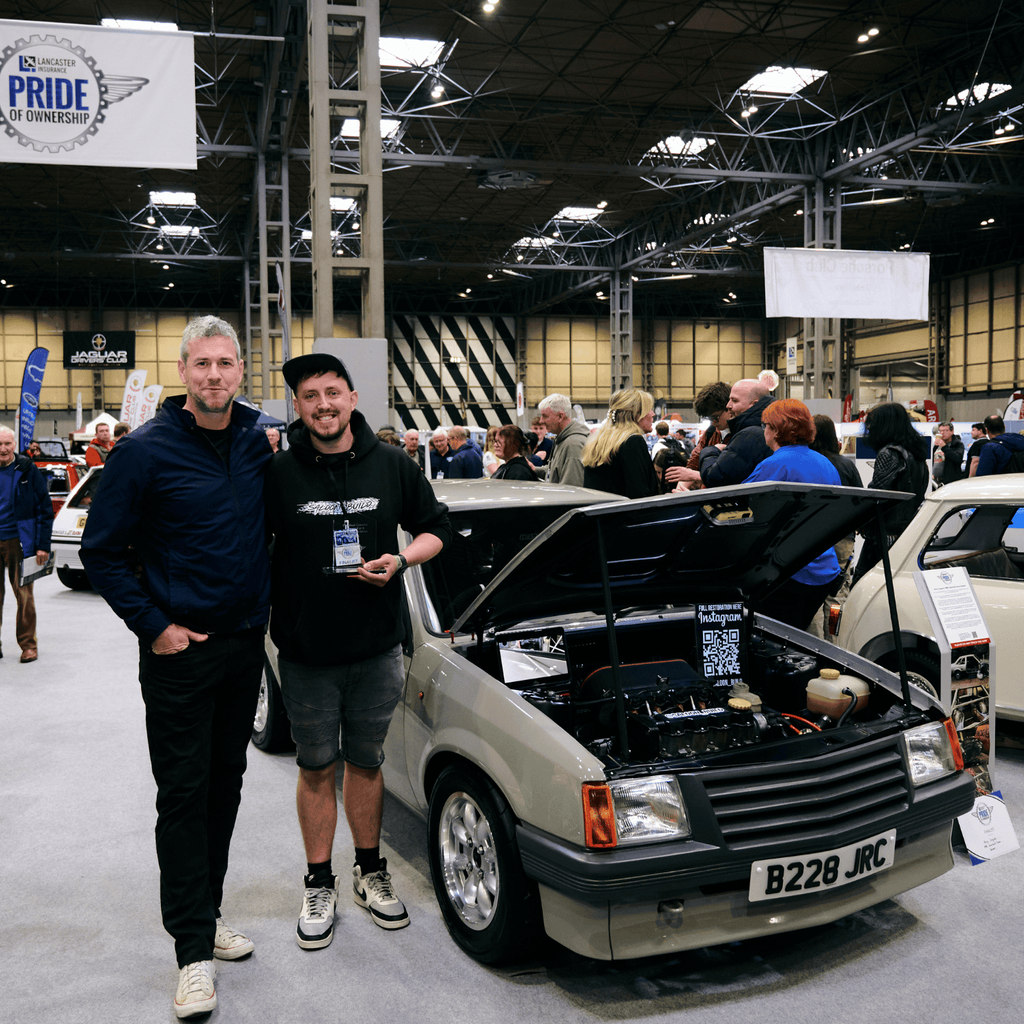 A light gray Vauxhall Nova Saloon is the main subject of this image, captured from a low angle at an indoor motor show. The vintage car is in excellent condition, with a clean design and sporty multi-spoke alloy wheels. A license plate is visible, reading 