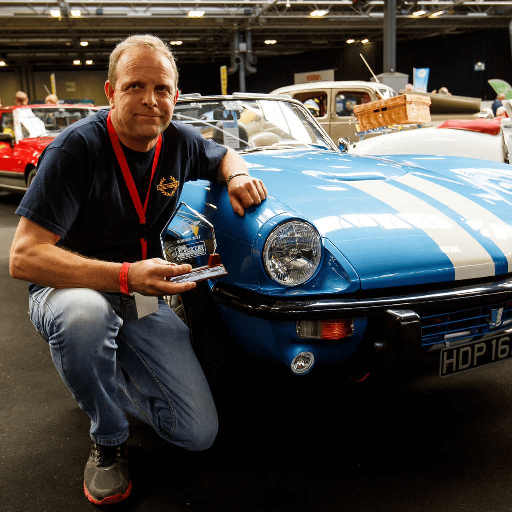 A man is crouching next to a blue Triumph Spitfire with white racing stripes at a car show. He is holding a trophy and smiling at the camera. The vintage convertible, which is the main focus, has a license plate that reads 