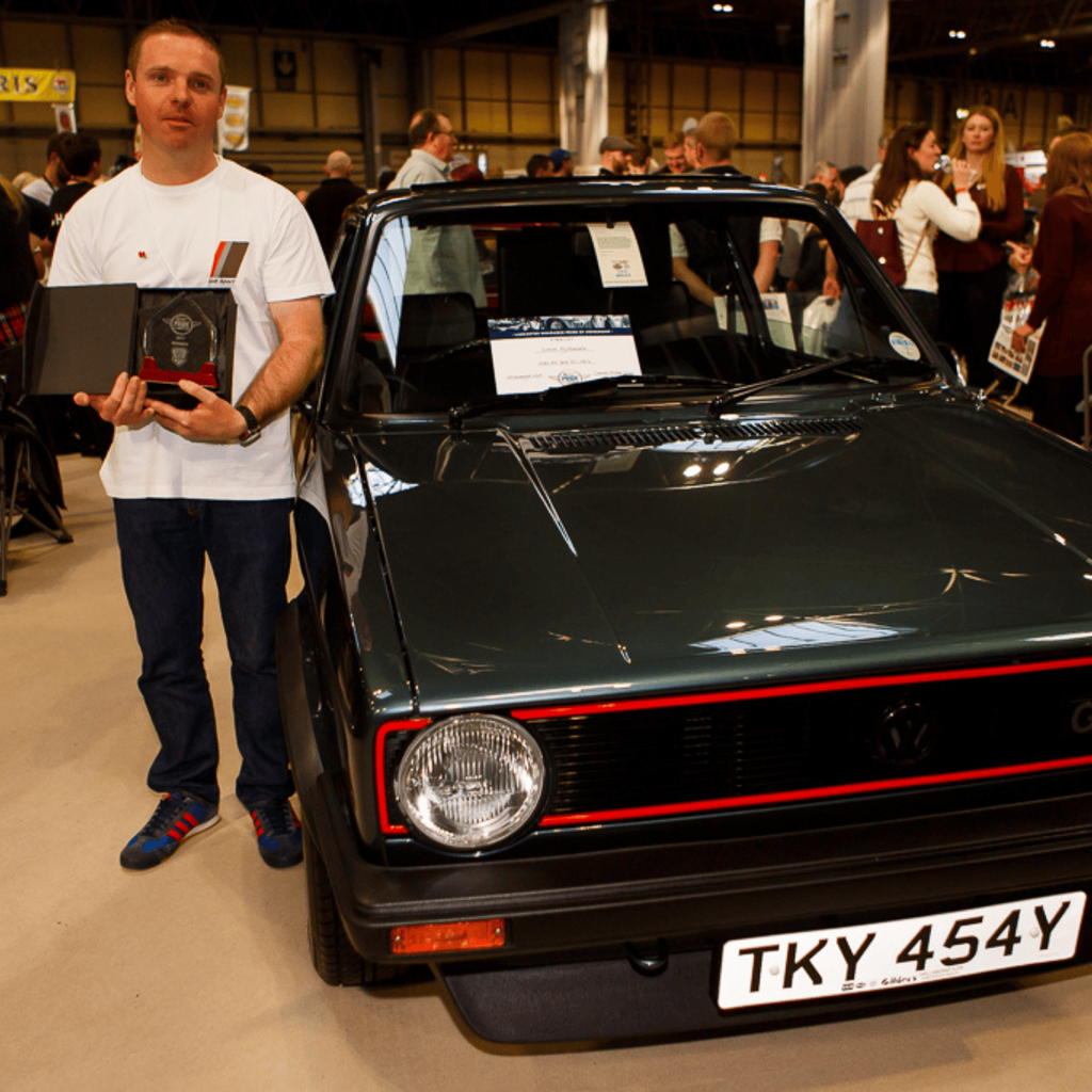 A man is standing proudly next to a vintage dark green Volkswagen Golf GTI Mk1 at an indoor car show. He is holding a trophy and a black display box, wearing a white T-shirt with the Audi Sport logo. The car, which is the main subject, has its iconic red grille stripe and the front license plate 