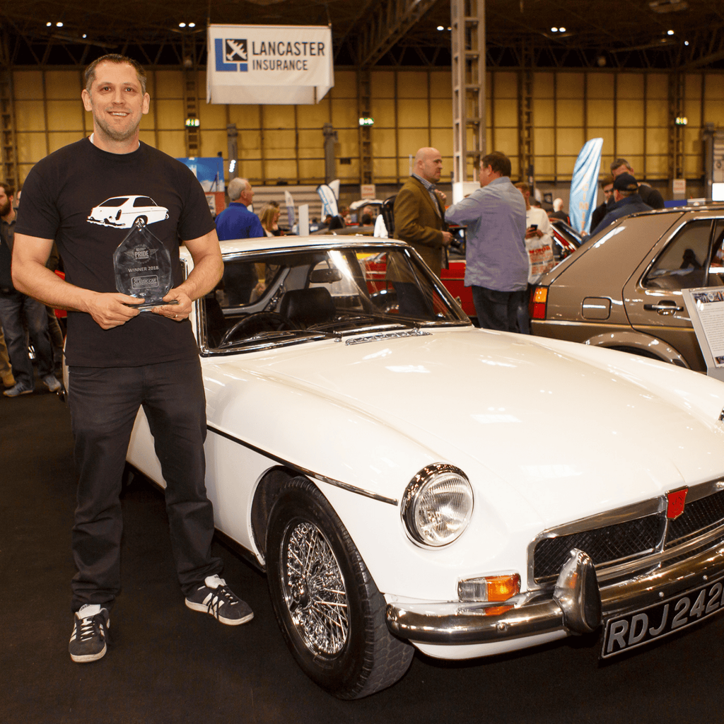 A proud man stands smiling next to a beautiful white MGB GT convertible at a car show. He is holding a trophy and is wearing a black T-shirt with an image of a car on it. The vintage British roadster, which is the main subject, is in excellent condition and features a chrome bumper and wire wheels. Its license plate 